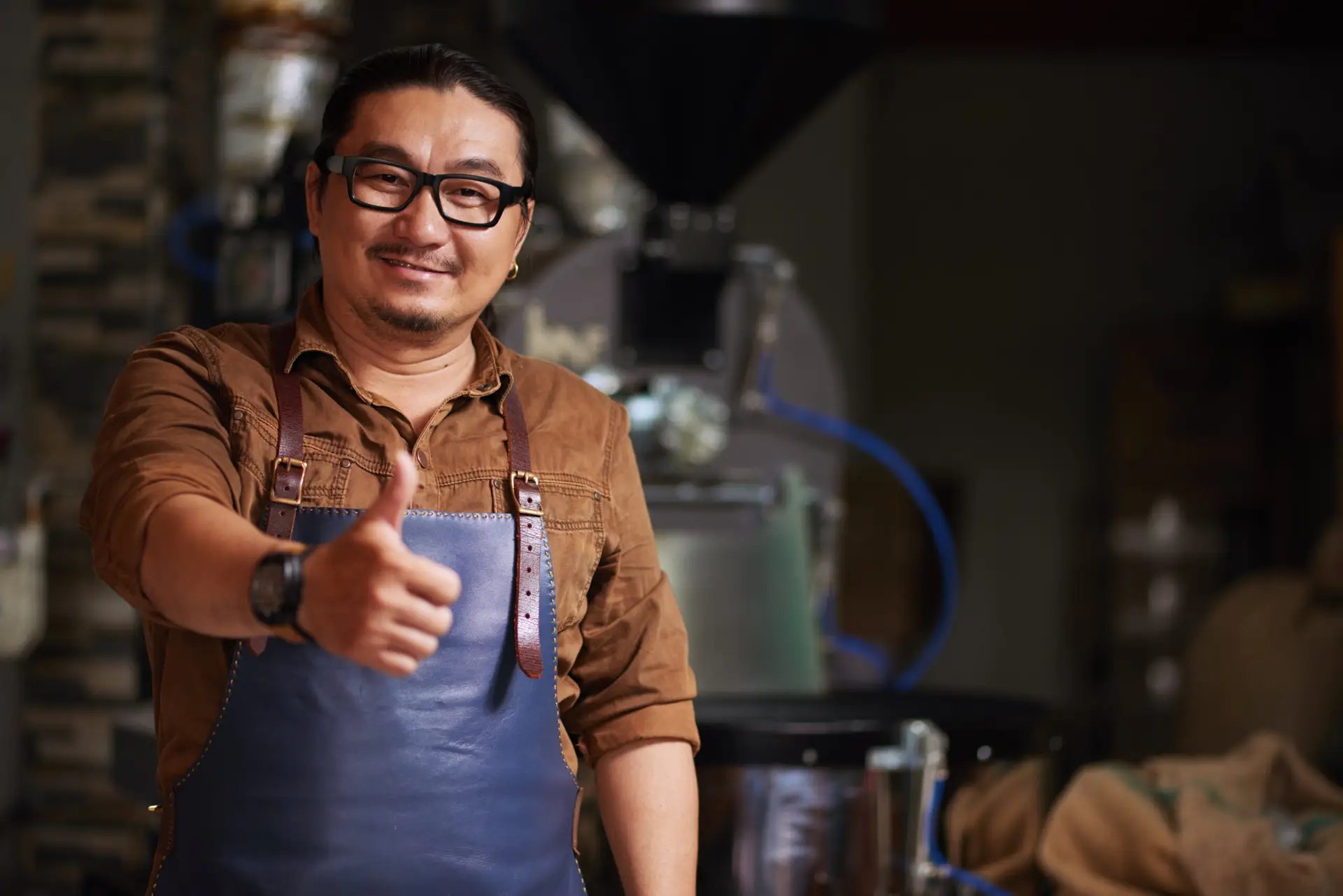 The cafe owner poses with a thumbs up in front of the coffee machine. He smiles happily because he has implemented proper small business management.